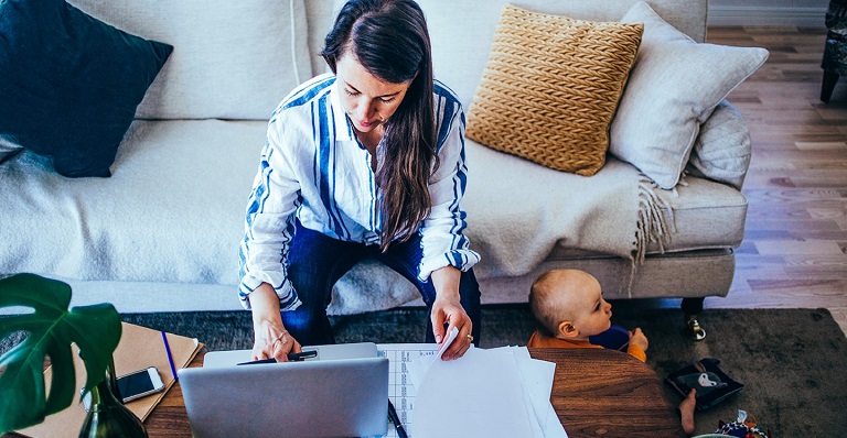 Woman sitting on the couch leaning over to her laptop with a baby sitting on the ground beside her