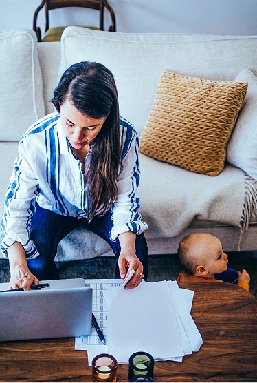 Woman sitting on the couch leaning over to her laptop with a baby sitting on the ground beside her