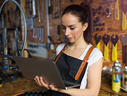 A young woman entrepreneur wearing overalls in a workbench environment stands looking at a laptop held in her hand.