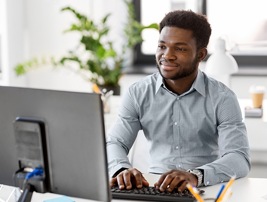 A young business owner wearing a button-down shirt types on a computer as he sits in a sunlit office.