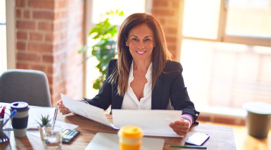 A woman smiling and sitting on a chair while reading documents at her desk.