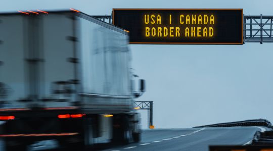 Blurry semi-truck approaching a highway sign for the U.S.-Canada border.