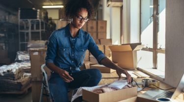 A woman seated on a step stool packing orders into cardboard boxes in an inventory room.