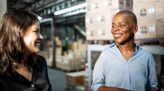 Two businesswomen walking and smiling in an inventory room.