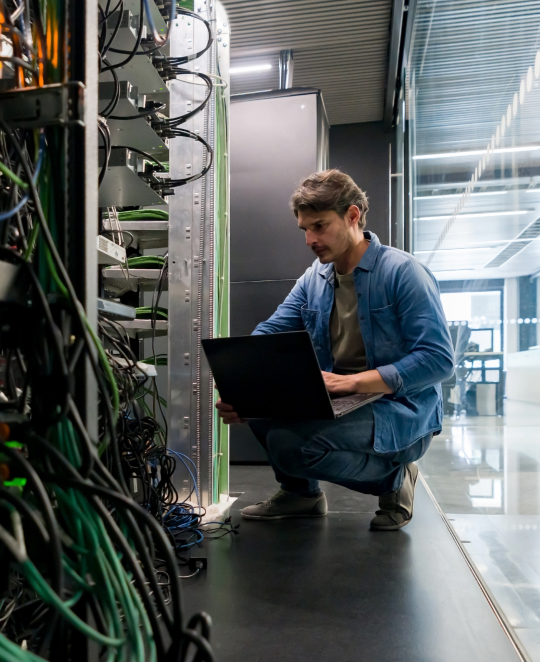 Person using a laptop while working beside server racks and network equipment in a data centre. 