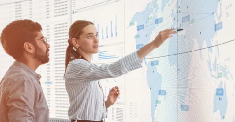 Two people reviewing global data visualizations and a world map on a large digital display.