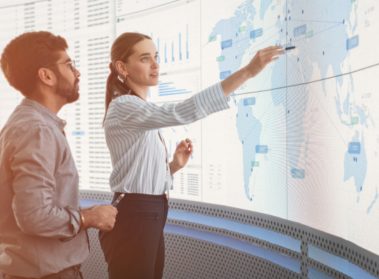 Two people reviewing global data visualizations and a world map on a large digital display.