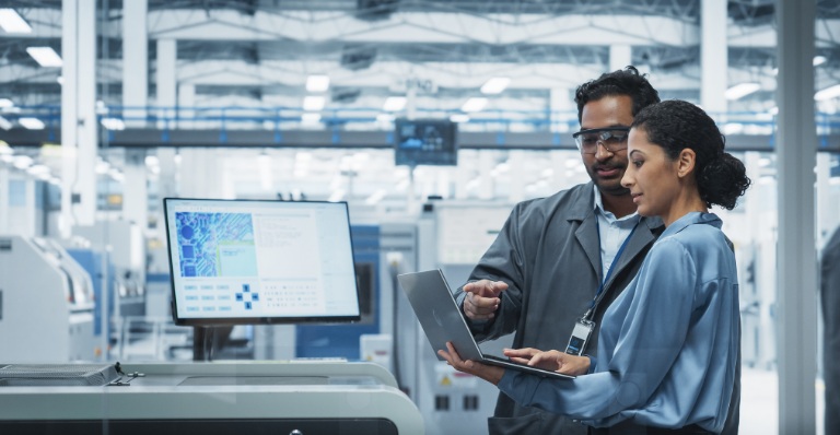 Two people reviewing information on a laptop beside industrial equipment in a manufacturing facility.