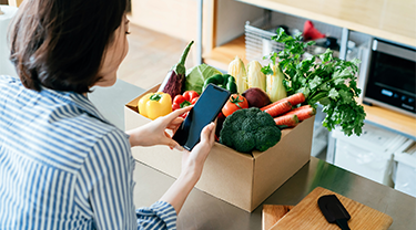 A woman with a mobile phone in her hand looks at a box of fresh produce on a kitchen counter. 