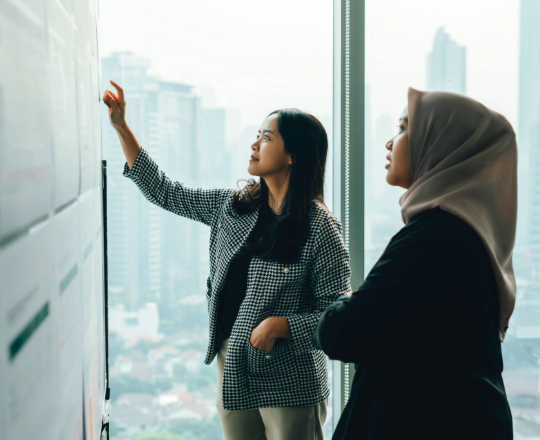Two businesswomen working and discussing ideas on a whiteboard in a modern office with a city skyline visible through the windows.
