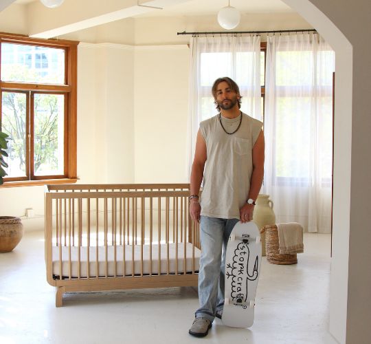Justin Segal in casual clothing holding a skateboard labeled “Storkcraft,” with a wooden baby crib in a nursery.