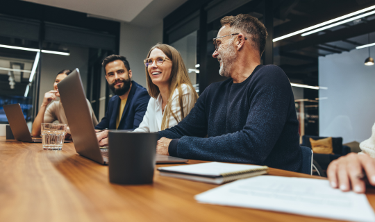 Four people sit in a modern meeting room at a wood table talking happily 