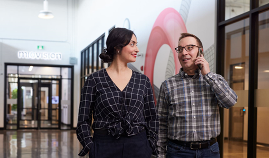 A man on a cellphone walks with a woman down a hall with windows