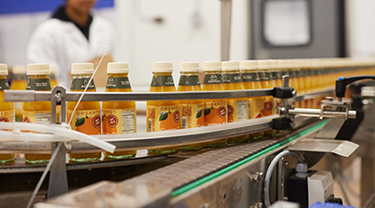 Bottles of organic orange juice on a conveyer belt. A woman in a white coat is in the background.  