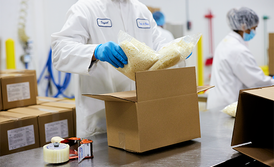 Bags of shredded cheese are loaded into a box by a man wearing blue gloves.