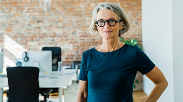 Une femme élégamment vêtue, avec des cheveux gris et des lunettes, se tient avec assurance dans un bureau.