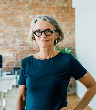 A smartly dressed woman with grey hair and glasses stands confidently in an office