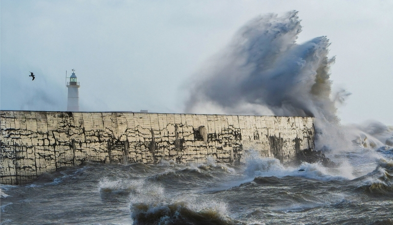 Waves crash against Newhaven Harbour breakwater during a storm, sending dramatic spray high into the air.