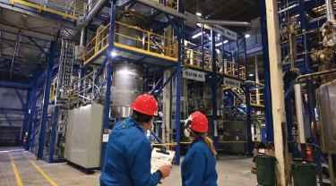 Two people stand in front of chemical processing machinery at GREENMANTRA. 