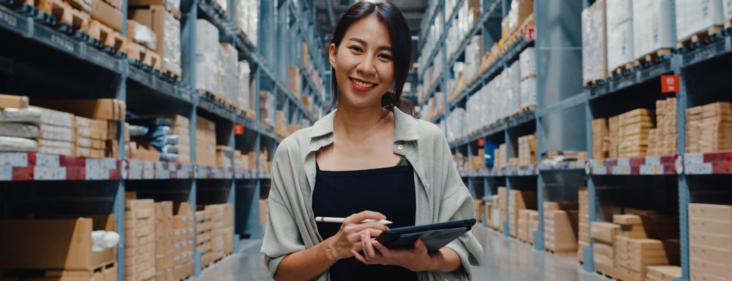 A woman wearing casual clothes, holding a tablet and tablet pen, smiling at the camera in an organized inventory room.