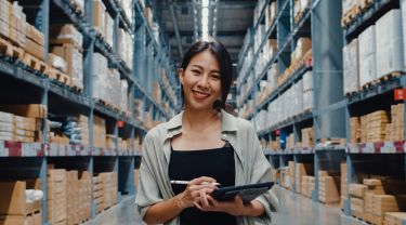 A woman wearing casual clothes, holding a tablet and tablet pen, smiling at the camera in an organized inventory room.