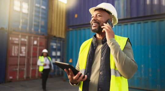 A smiling man in a safety vest and hat talking on the phone while holding a tablet in a shipyard, with a blurry woman in the background also wearing a safety vest and hat.