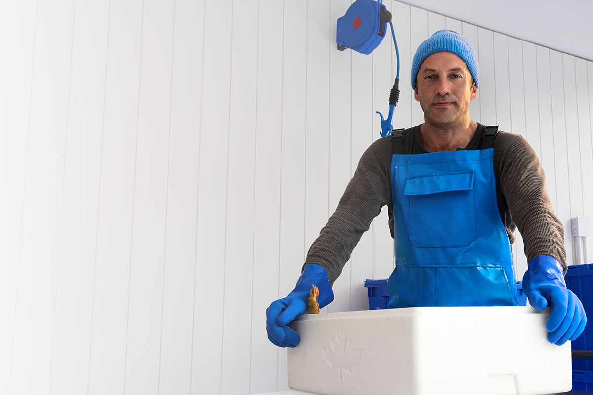 Un pêcheur portant une salopette bleue et des gants se prépare à livrer des fruits de mer dans des contenants bleus, ornés d’une feuille d’érable.