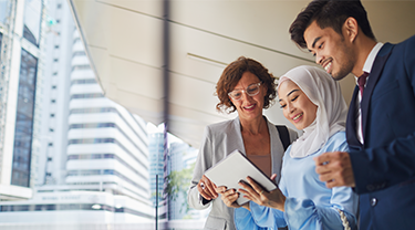 A woman with curly hair and glasses, a woman wearing a white hijab and a man in a blue suit look at a digital tablet while standing on a balcony.