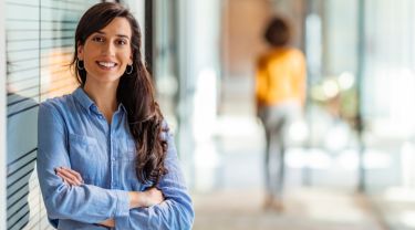 A businesswoman leaning against a glass wall in an office with another woman walking away in the blurred background.