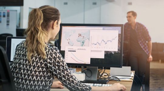 A businesswoman at a desk scrolling on a computer displaying a map with data points and graphs, with a blurred businessman in the background of an office.