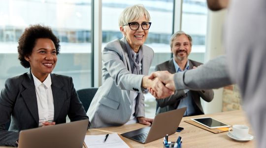 A woman smiling and shaking hands with a man, while a smiling woman sits to her left and a man claps to her right.
