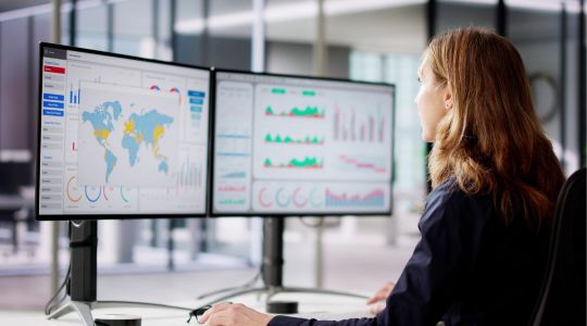 A businesswoman at a desk viewing maps and data on dual monitors in an office setting.