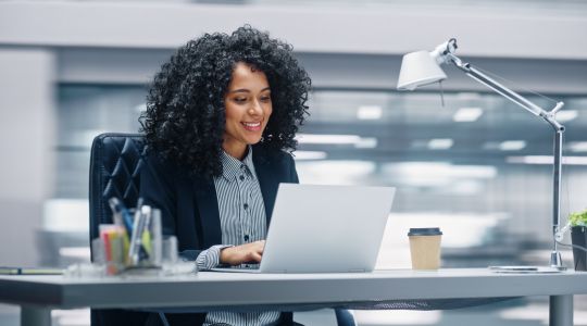 A smiling businesswoman sitting at a desk, working on her laptop.