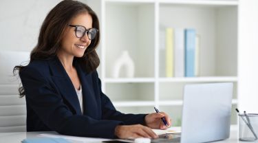 A smiling businesswoman wearing glasses working on a laptop at a desk in a modern office.