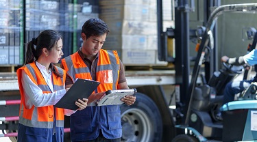 Warehouse workers double-check paperwork as forklift loads shipment into a truck.