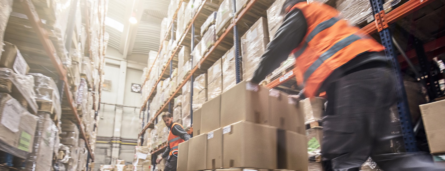 Two workers move large pallet of boxes through a warehouse.