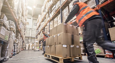 Two workers move large pallet of boxes through a warehouse.