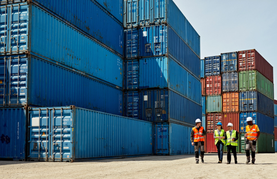 A group of workers in safety vests and hard hats standing next to a large stack of shipping containers