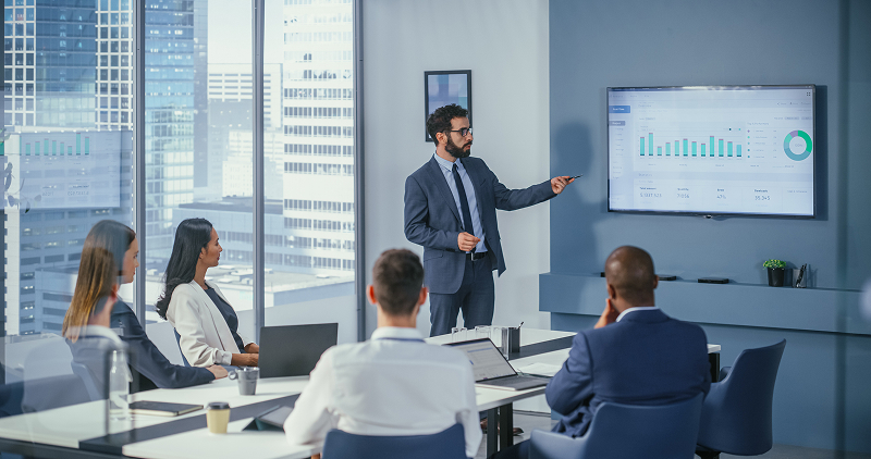 Person presenting an investment strategy in a conference room with data and stock market trends on a wall screen.