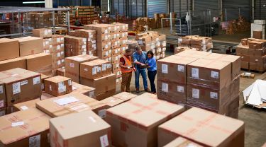 Two men, one in a safety vest with gloves holding a scanner, the other casually dressed, and a woman in business casual holding a tablet, in a large warehouse full of boxes.