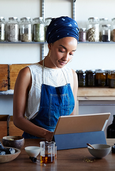 A woman working on her laptop computer while standing in an organic products workspace surrounded by jars of ingredients. 