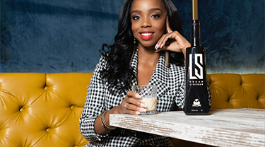Myriam Jean-Baptiste sitting at a table holding a drink, a bottle of LS Cream in front of her 