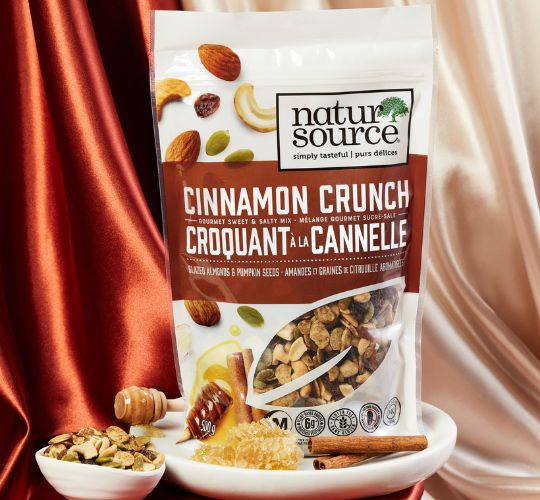A bag of naturSource cinnamon crunch mixed nuts on a plate with cinnamon sticks, a honeycomb and a honey stick. A bowl of mixed nuts is on the side, with silky red and light pink drapes in the background. 