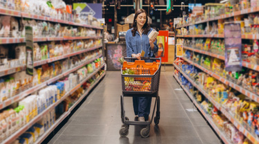 Woman standing in a grocery store aisle, holding a product and pushing a shopping cart.