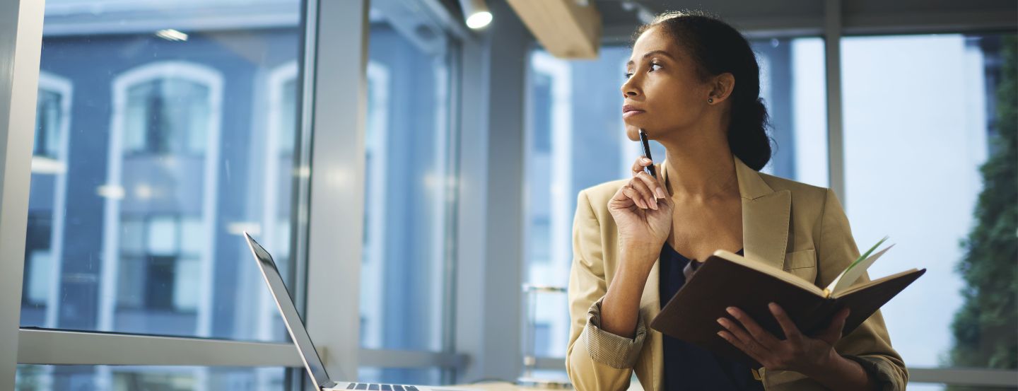 Une femme d'affaires tenant un cahier et un stylo, regardant par la fenêtre d'un bureau.