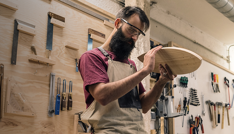 A man works on a piece of wood in a woodworking shop surrounded by his tools and carpentry square.