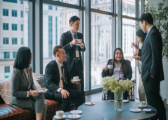 A group of six businesspeople drink coffee together around a table in an office meeting room.