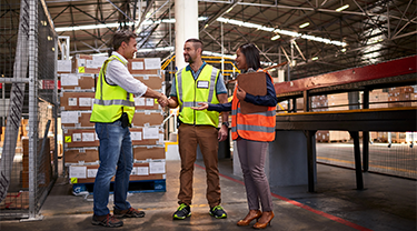 Two male workers shake hands in a warehouse, while a third female worker looks on 