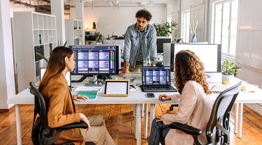 Three co-workers (one male and two females) look at colourful graphs on their computer screens.