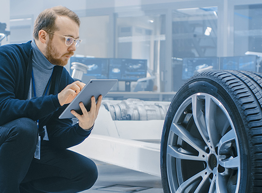 Man holding tablet inspects an automobile frame and wheel assembly in a factory.